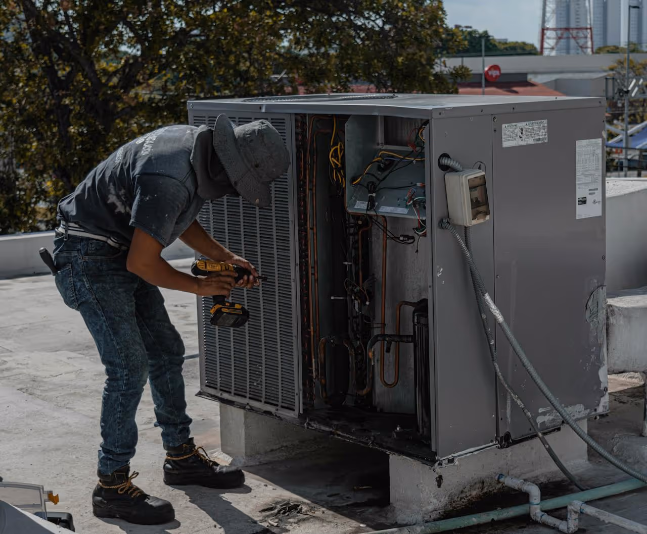 Technician wearing a hat and boots repairing an outdoor HVAC unit with a cordless drill on a rooftop.