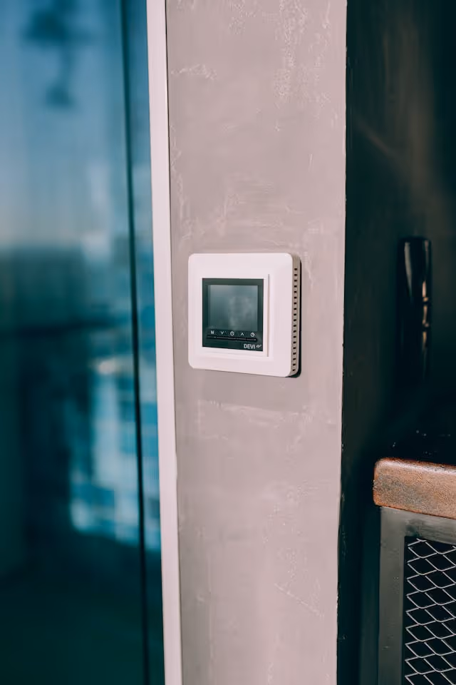 White digital thermostat mounted on a textured beige wall next to a glass door and a countertop.